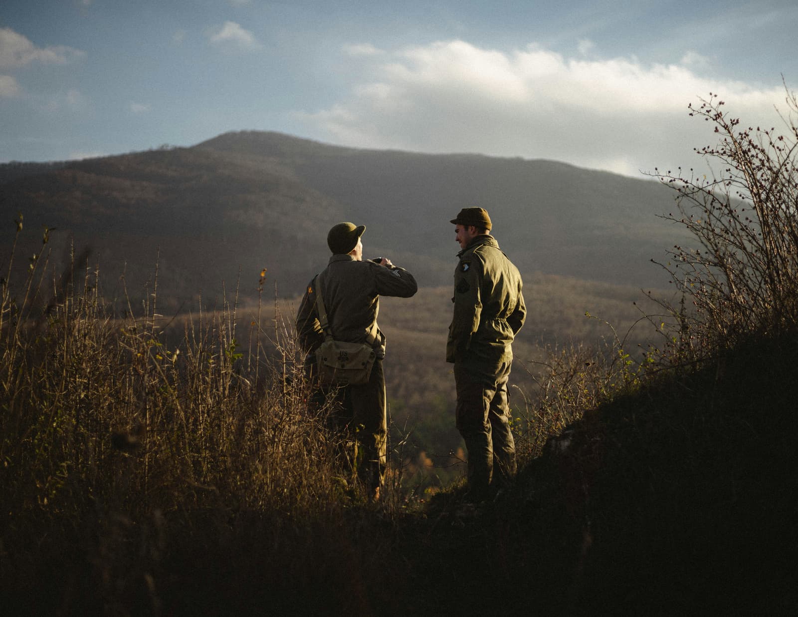 Two hunters standing on a hillside overlooking a mountain valley at golden hour