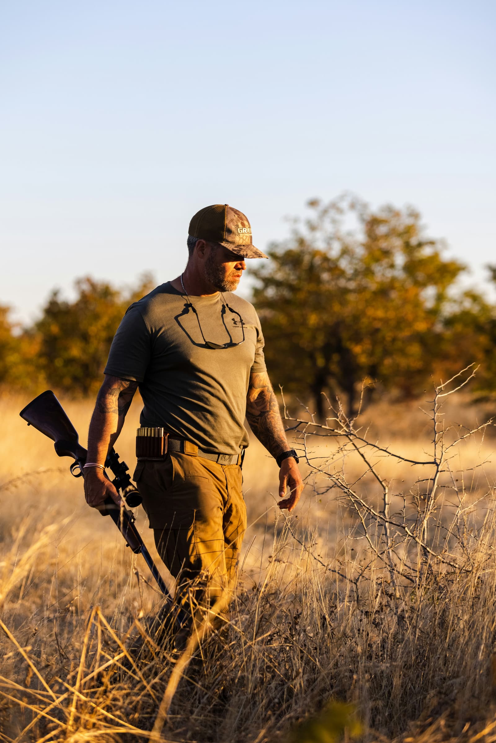 Hunter walking through dry grassland with rifle at golden hour